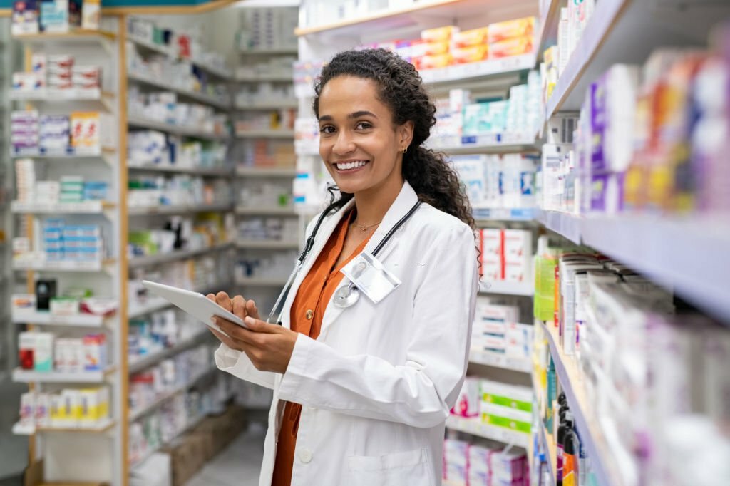 Happy friendly multiethnic pharmacist doing inventory in a provided and modern pharmacy while looking at camera. Portrait of smiling young doctor woman working in drugstore with digital tablet. African smiling druggist working at hospital pharmacy.